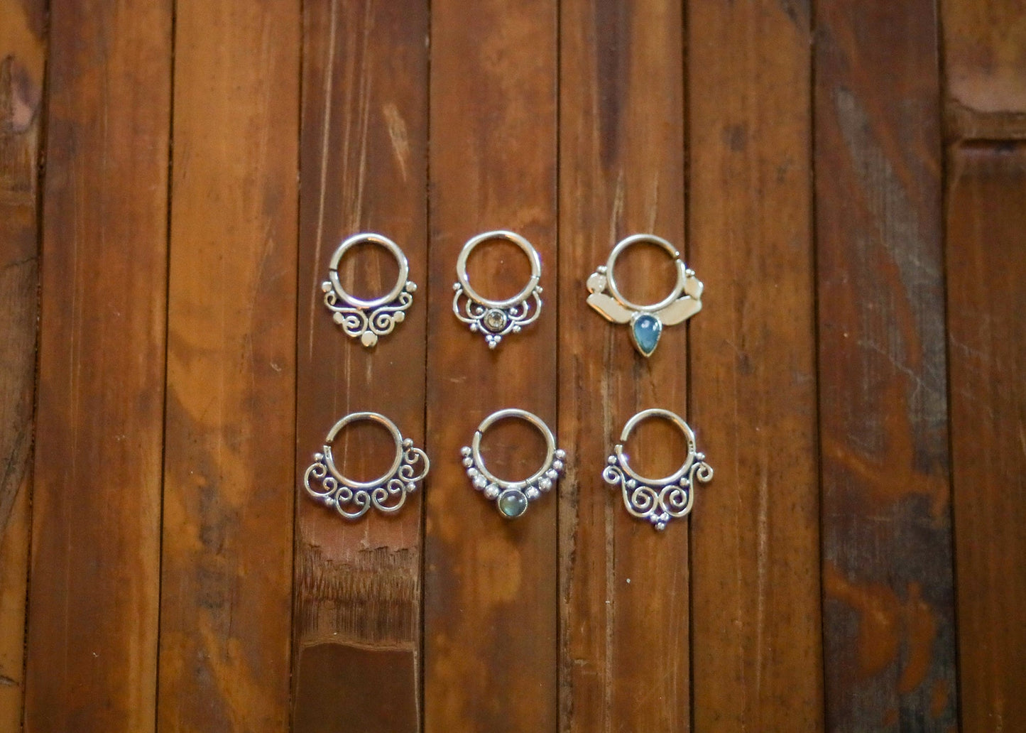 May include: Six silver septum rings with decorative designs. Three rings have a small blue gemstone in the center. The rings are displayed on a wooden surface.